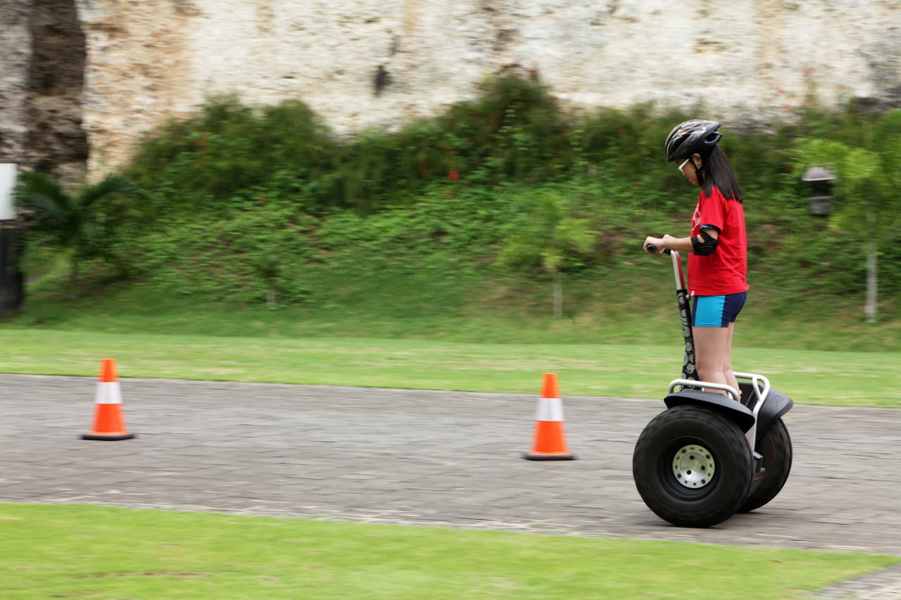 A young girl in safety gear riding a Segway on a paved path outdoors, surrounded by nature.